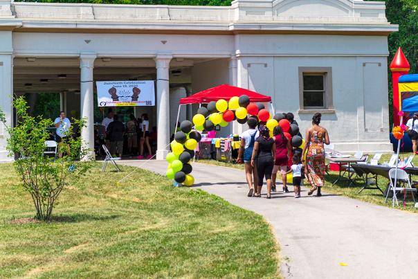 A group of people walk under a balloon arch up to a park pavilion to celebrate a festival