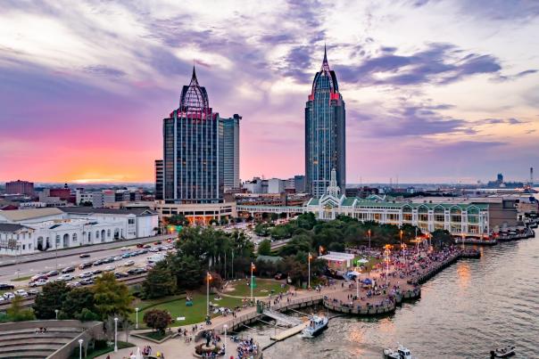 A vibrant sunset over Downtown Mobile skyline, featuring tall buildings, a bustling waterfront, and a lively crowd below.