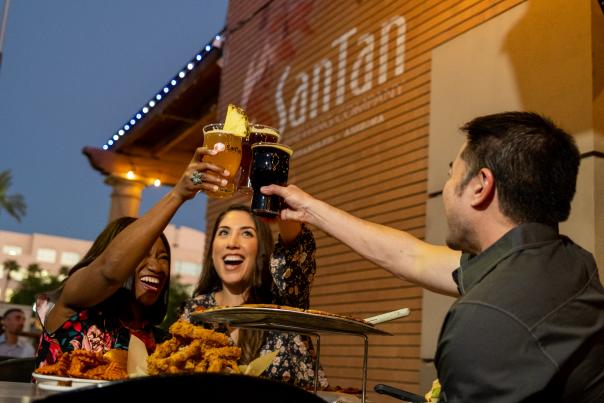 Two women and one man cheers beers on SanTan Brewing patio with food.