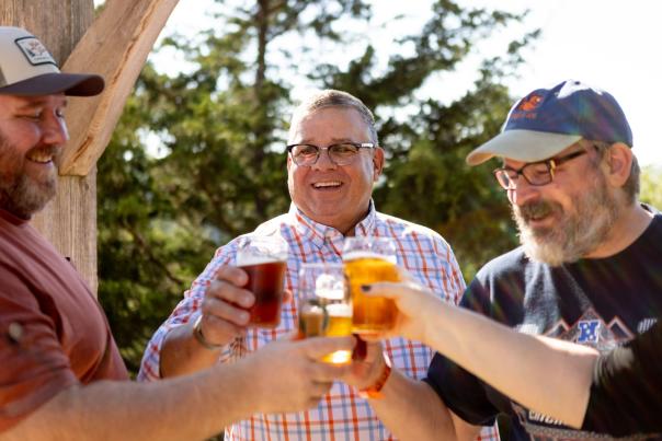 A group of three white men cheers beers while standing outside at New Glarus Brewing Co.