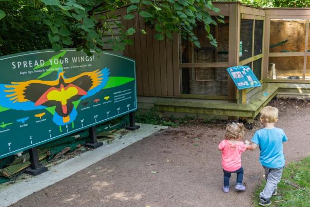 Chris Ahern Photography, Chris stands outside County Farm Park under the 'County Farm Park' archway, with one child in a stroller and one child holding Chris' left hand. Green trees line the background.