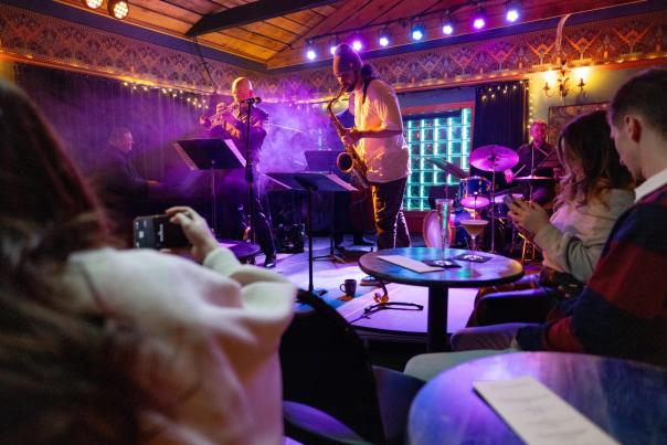 A man playing the saxophone along with other musicians perform on a stage in a dimly lit jazz club as the audience enjoys cocktails.
