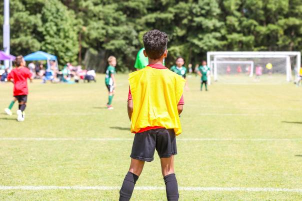 A child wearing a yellow practice vest stands on a grassy soccer field and looks toward a youth match in progress, with teammates, goals, and tree-lined surroundings visible ahead.