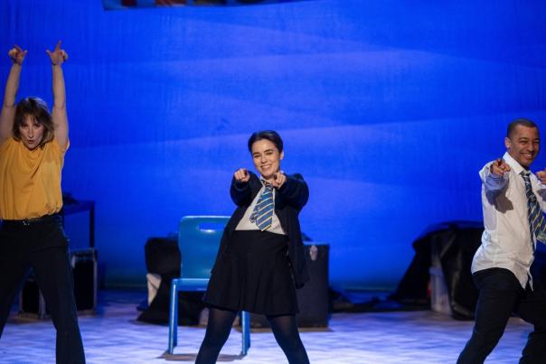 A still from John Godber's play 'Teechers'. Two actresses and an actor stand on stage dressed in school uniform with ties and shirts. They smile and point towards the photographer.