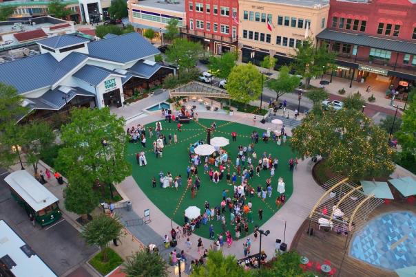 The drone camera overlooks Market Street's Sixty Vines and Central Park. The park is studded with fancy white sun-umbrellas and well-dressed people relaxing after the TSAE conference sessions. There's a town trolley featured in the lower left and the splash pads in the lower right.