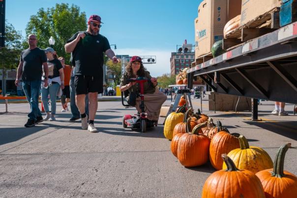 A street fair scene with pumpkins lined up on the ground, people walking by, including one person on a mobility scooter. The atmosphere is lively and sunny.