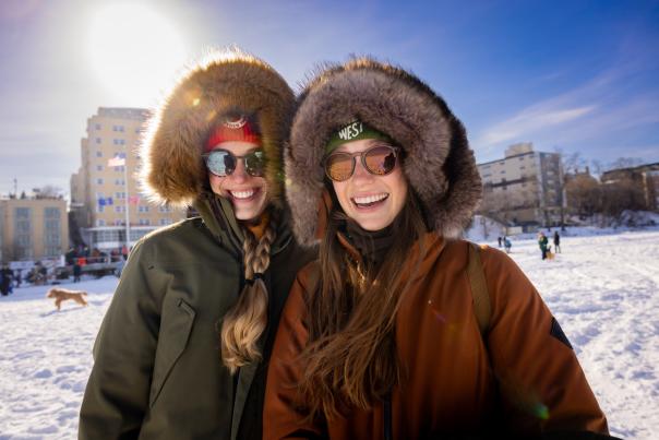 Two white women in coats with fuzzy hoods smile while standing on a frozen lake in front of the Edgewater Hotel