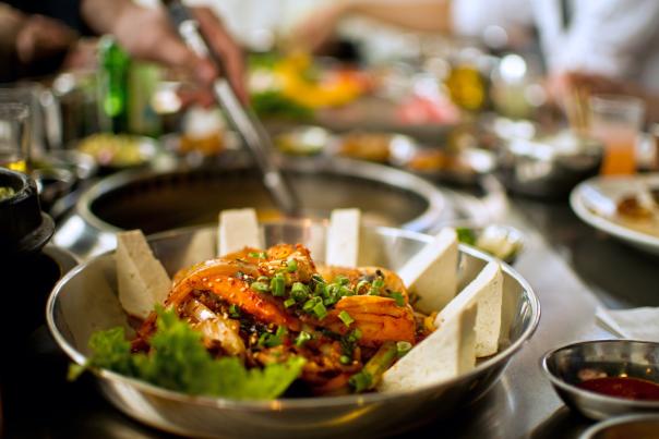 A prepared dish sits ready to be served in a steel bowl as someone's hand reaches in for a serving on a separate dish in the background.