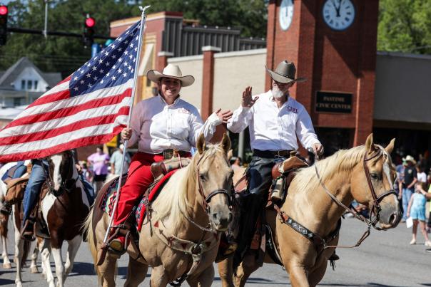 A man and a woman holding an American flag are riding horses in the street for a parade.