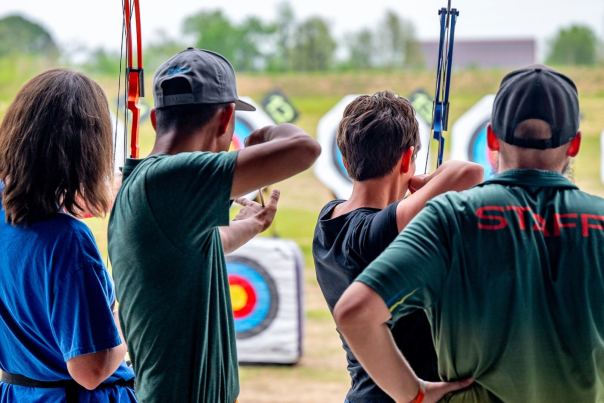 The image shows four individuals at The Quiver Archery Range in Osage Park, Bentonville, Arkansas, practicing archery and aiming at colorful targets.