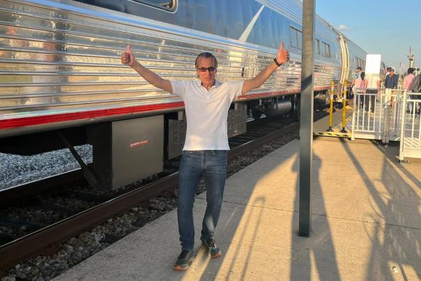 A man in sunglasses, a white shirt and jeans holds two thumbs-up while standing in front of a passenger train