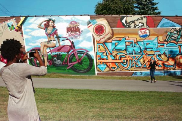 A woman photographs her friend posing for a photo in front of a series of huge, vibrant murals.