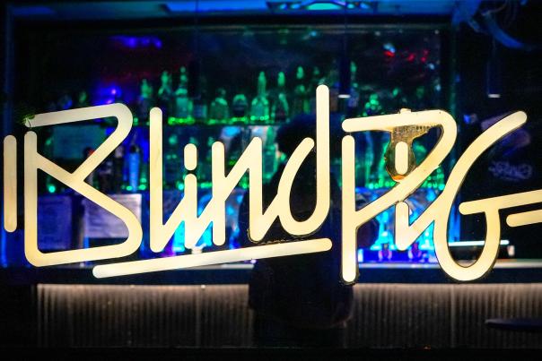 Window cling that reads "Blind Pig". Inside of the glass, a person stands at a dimly-lit bar.