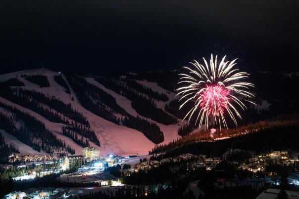 Fireworks illuminate a snowy mountain landscape at night, with a ski resort and cozy lodges nestled in the valley below.