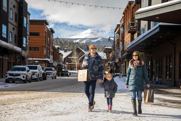 A family walks along a snowy street lined with shops, with a mountain backdrop. The father carries a shopping bag, while the mother and child enjoy the day.