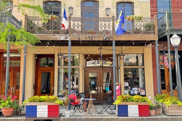 A two-story building with a wrought iron balcony is decorated with French flags. There are tables and chairs on the sidewalk.