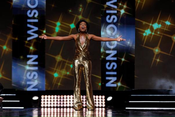 Willow Newell in a gold jumpsuit singing on stage with Miss Wisconsin on the screens behind her