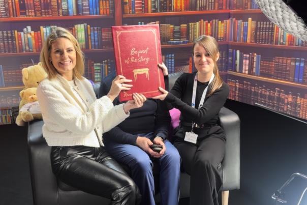 Two women sit on a black sofa holding up a red book which says Be Part of the Story in gold letters. The book cover hides a third person's face who is sat in the middle of the sofa. They are in a film studio with books and bookcase imagery projected onto screens around them.