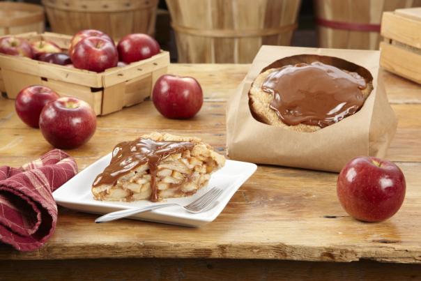 Slices of caramel-covered apple pie served on a white plate next to a whole pie in a paper bag, surrounded by fresh red apples and rustic wooden baskets.