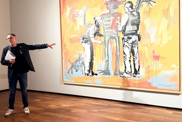 An art museum curator wearing jeans and a black jacket point to a large painting in an art museum