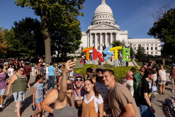 A group of six white adult men and women take a selfie in front of a balloon installation that says TASTE at the Wisconsin State Capitol during Taste of Madison