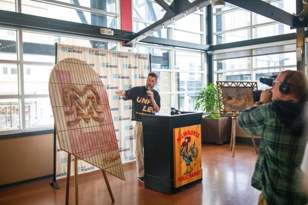 A speaker presents a wooden art piece featuring the Milwaukee ‘M’ symbol during a press event at the Milwaukee Public Market, with a Visit Milwaukee backdrop and a camera operator filming the moment.