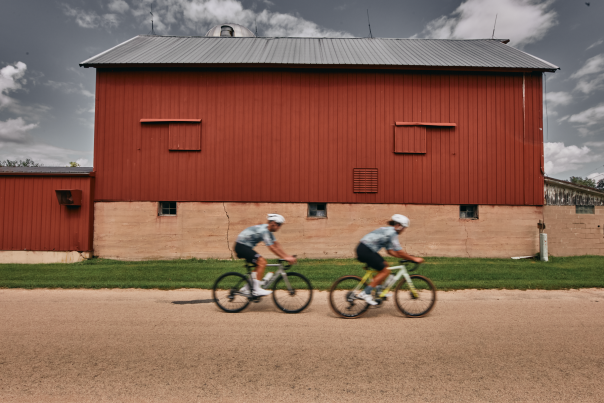 Two cyclists riding in front of a red barn.