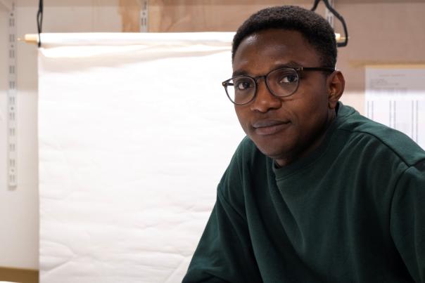 Ghanian artist, Isaac Yeboah sits in a green jumper wearing glasses. He looks towards the camera and sits a desk with boards in the background leant against a wall. He sits in an art studio.