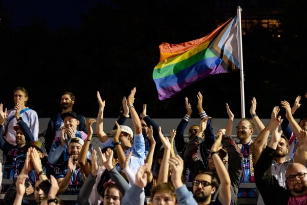 Pride flag waves above fans with arms raised at a Forward Madison FC soccer game