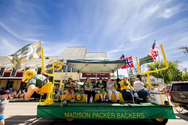 A green and yellow float that says Madison Packers Backers with several people sitting on it rides down the street during the Syttende Mai parade