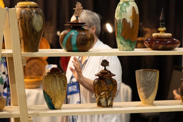 A man with gray hair stands behind a wood display at The Woodlands Fine Craft Show in 2025. Two shelves support a collection of vases and urns, each made of a different wood combined with colorful inlays.