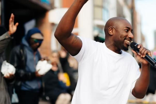 A man raises his arm with a microphone in the street and smiles with rappers behind him.