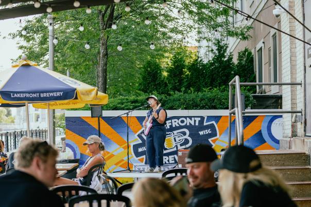 A woman in a navy blue romper singing into a microphone on a stage, holding a pink guitar, with people in front of her sitting at tables under gold and blue umbrellas on a patio along the riverfront with trees in the background.