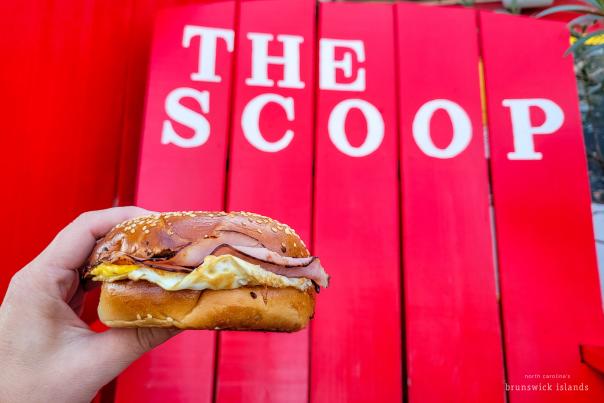 A hand holding an egg and ham bagel in front of a large chair