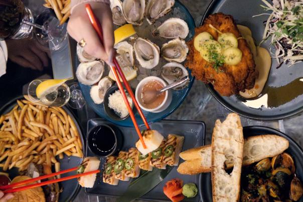 Overhead view of a vibrant and diverse group dinner spread at Bernie's Supper Club. Diners are reaching in with red chopsticks to share a variety of dishes, including oysters on the half shell, sushi rolls topped with jalapeños, fried chicken with pickle slices and slaw, crusty bread with mussels, a burger with fries, a noodle dish, and assorted dipping sauces. Wine glasses are also visible, creating a lively, upscale dining atmosphere.