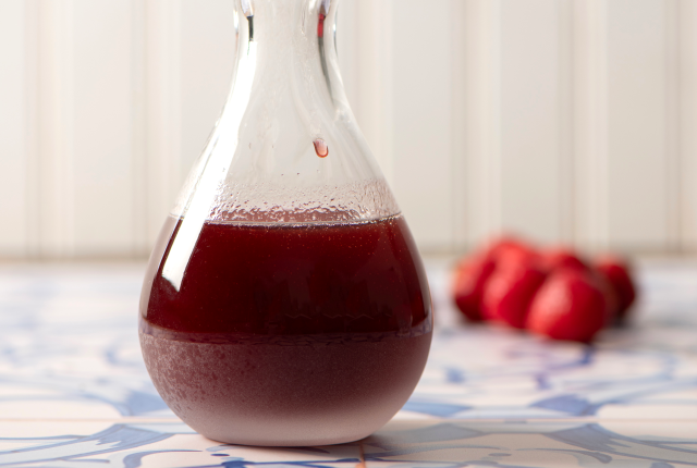 Glass container filled with red prickly pear syrup with prickly pears in the background on a white and blue table.