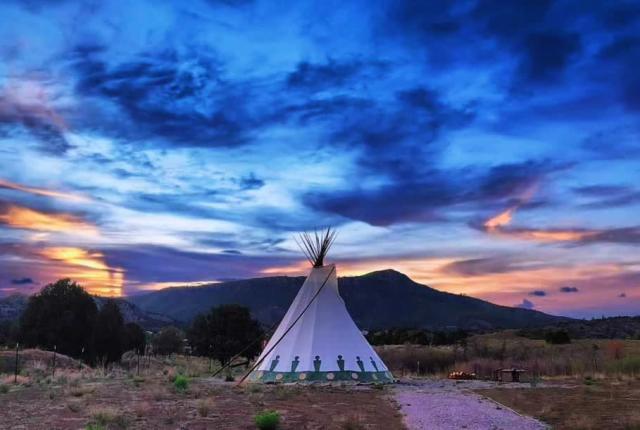 A teepee stands in a serene landscape under a vibrant sunset sky with pink, orange, and blue hues.