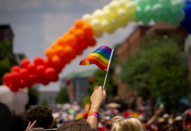 A person holds a rainbow flag beneath a rainbow balloon arch.