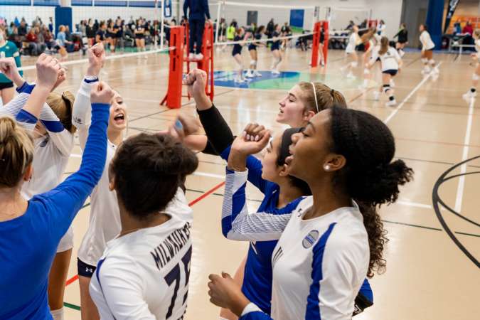 Five girls in volleyball uniforms stand in a ground to cheer on their team before playing a match. Spectators and the other teams are visible in the background.