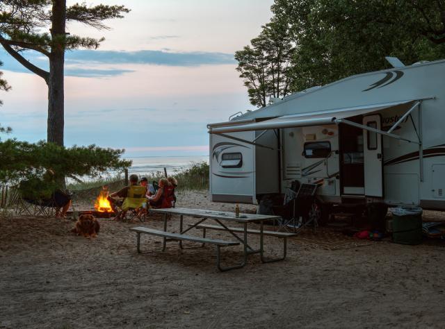 A family sits by a fire pit alongside their white camper at Southwick Beach State Park