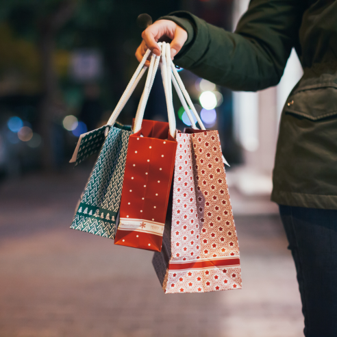 A woman wears a green jacket and holds up three red, white, and green Christmas shopping bags of different sizes. The background is blurred.