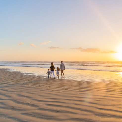 A family of four walks on the beach away from the camera towards the left. The sun is rising to the right.