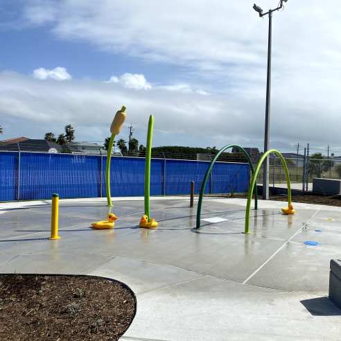 On a gray pad sits several green and yellow hoops and poles with water for kids to splash on. A playground is in the back right.
