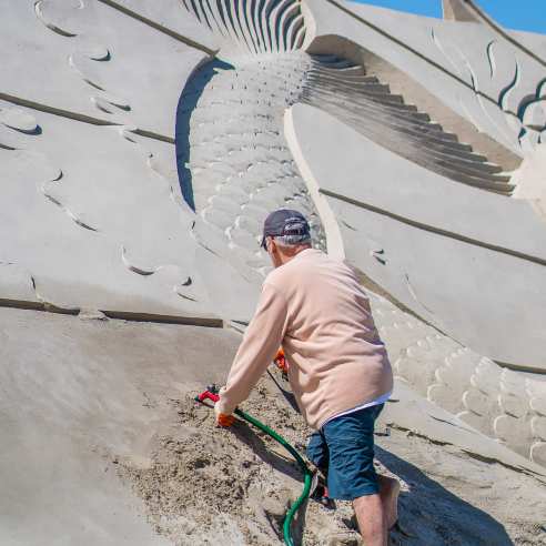 A man in an orange, long-sleeve shirt and blue cutoffs uses a hose and sculpts on a massive sand sculpture. A mermaid tail is visible on the mountain.