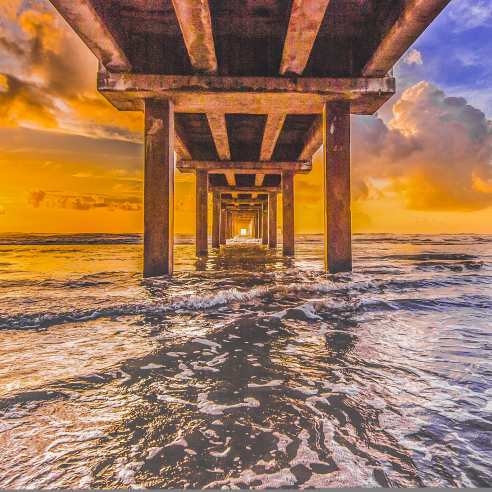 The sun rises just over the water from the perspective of under a pier