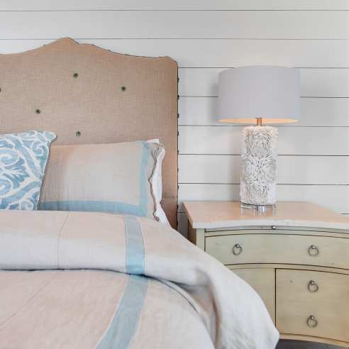 Image of a tan bed and nightstand against a white shiplap wall. On top of the nightstand is a shell lamp.