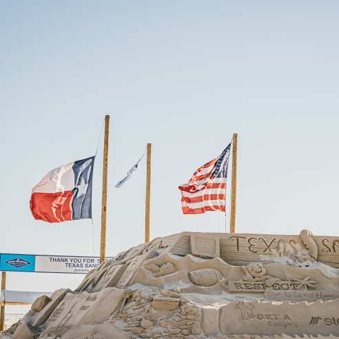Three flag poles with the US flag, Texas flag, and a Texas SandFest flag fly on poles behind a large sand mountain carved with sponsor logos