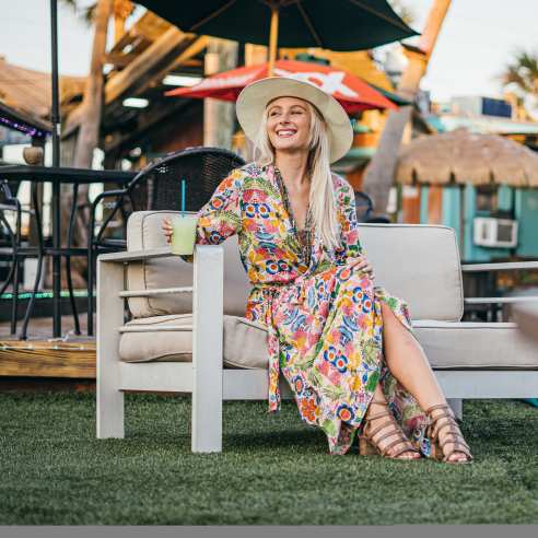 A girl in a springy floral dress and a hat sits on a couch in an outdoor green space. She holds a drink in one hand and smiles off camera.