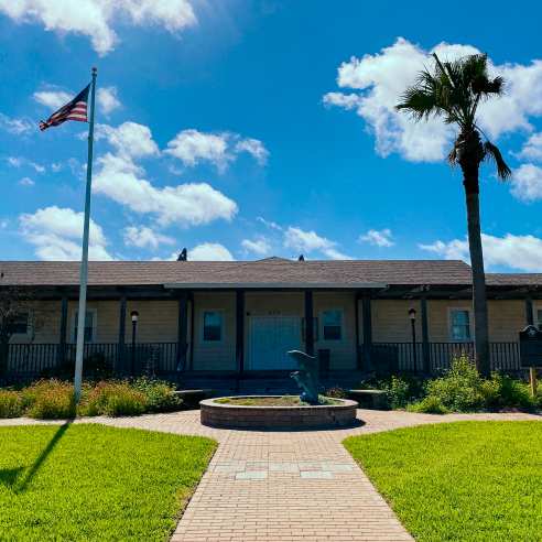 A long low yellow building sits at the end of a pathway surrounded by grass. A blue sky overhead.