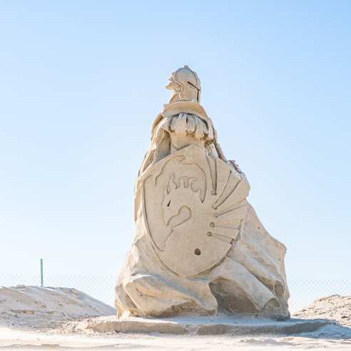 Sand sculpture on the beach of a guardian soldier facing the left. The soldier carries a shield emblazoned with a dragon.
